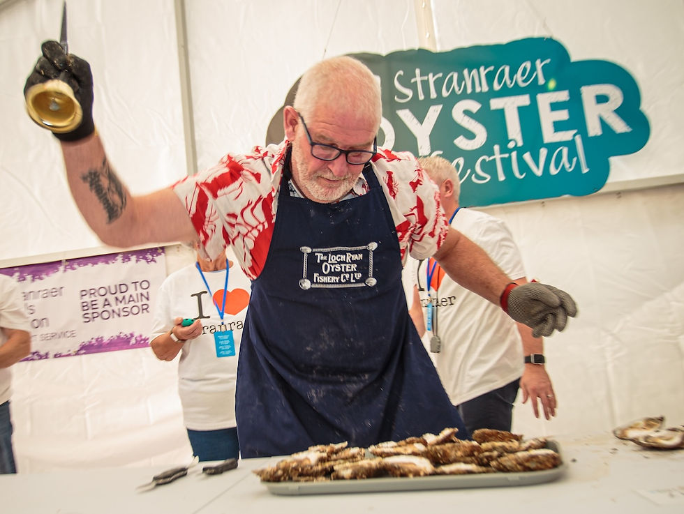 Man in apron shucks oysters at Stranraer Oyster Festival. Others in branded shirts watch. Vibrant, lively atmosphere, event signage visible.