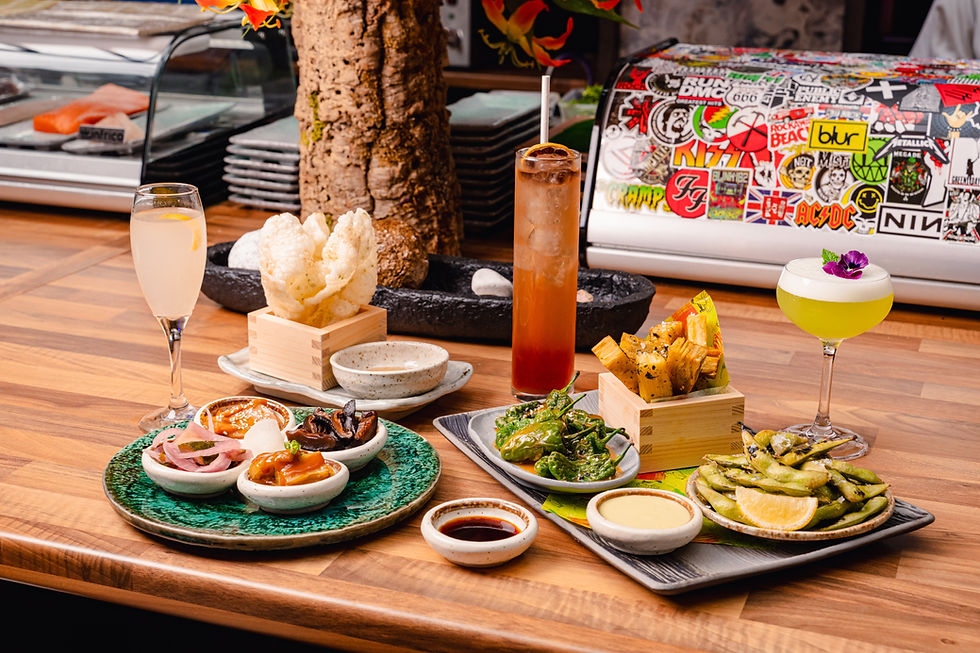 A vibrant spread of appetizers, drinks, and sauces on a wooden table. Background features a sticker-covered sushi case and decor.