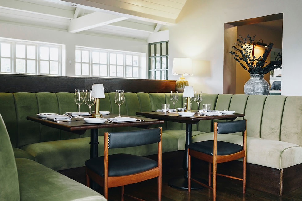 Empty restaurant booth with green velvet seating, set tables and wine glasses, warm lamp, and a large vase of branches in the background