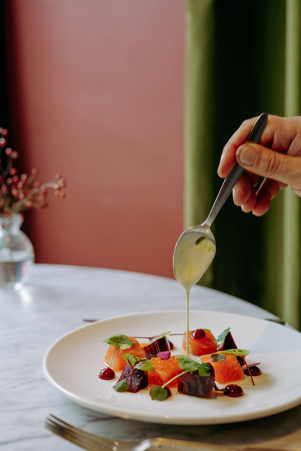 Hand pouring sauce over a gourmet salmon and beet dish on a white plate, with a pink and green restaurant backdrop.