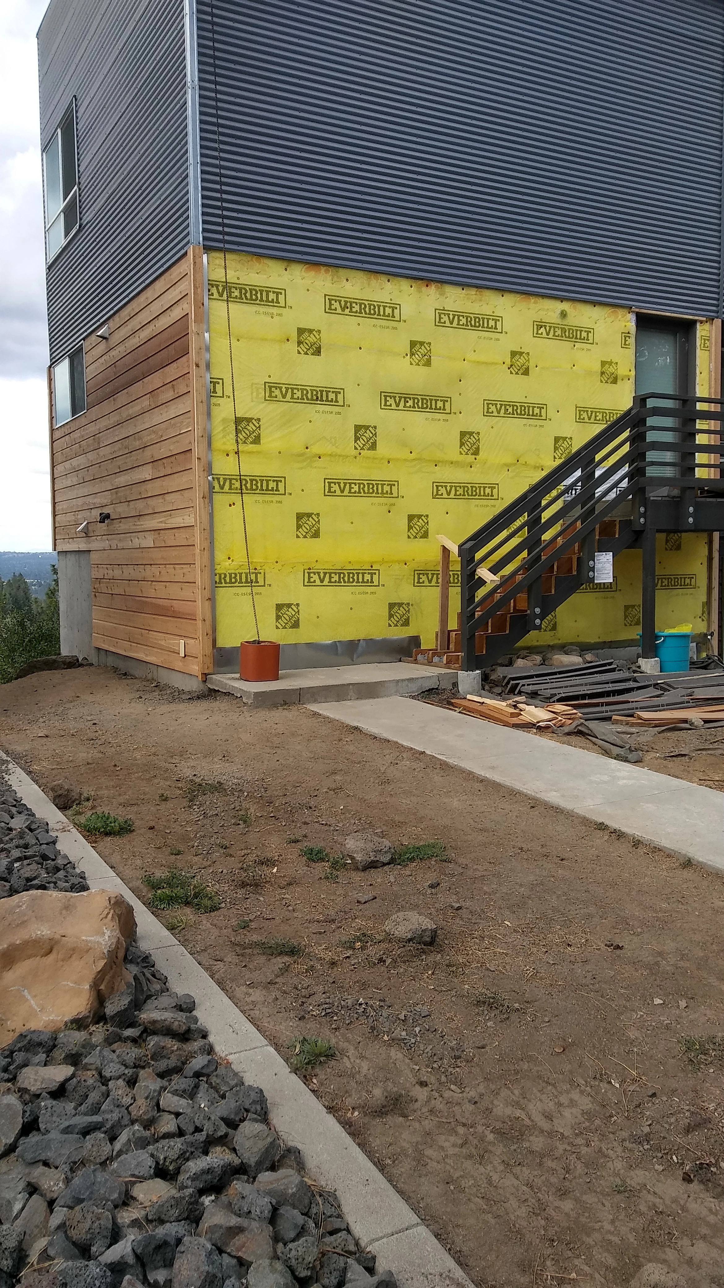 Expert siding repair and installation in progress on a stunning mountain-side home in Spokane Valley and Spokane, WA. This image showcases our process, with yellow rain screen mesh applied over a white Tyvek vapor barrier on one side and completed Channel wood siding without exterior stain on the other. The stunning mountain backdrop and newly landscaped surroundings add to the beauty of our work in progress.
