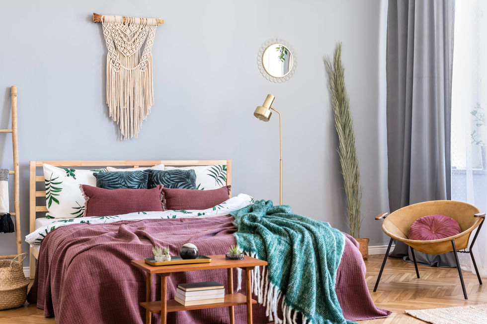 Bedroom with a macrame wall hanging, burgundy and teal bedding with botanical print pillows, a brass floor lamp, and a round mirror