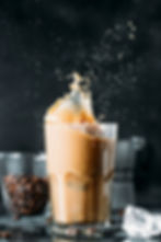 Cold iced coffee drink with milk and liquid. Ice cubes in tall glass mugs. Dark background with copy space. Refreshing cappuccino or milkshake design. Close up of cold iced drink with ice. Refreshing drink in late autumn. Glass of cold beverage with milk spilling out. Ice and chocolate bar, liquor on a wooden background.