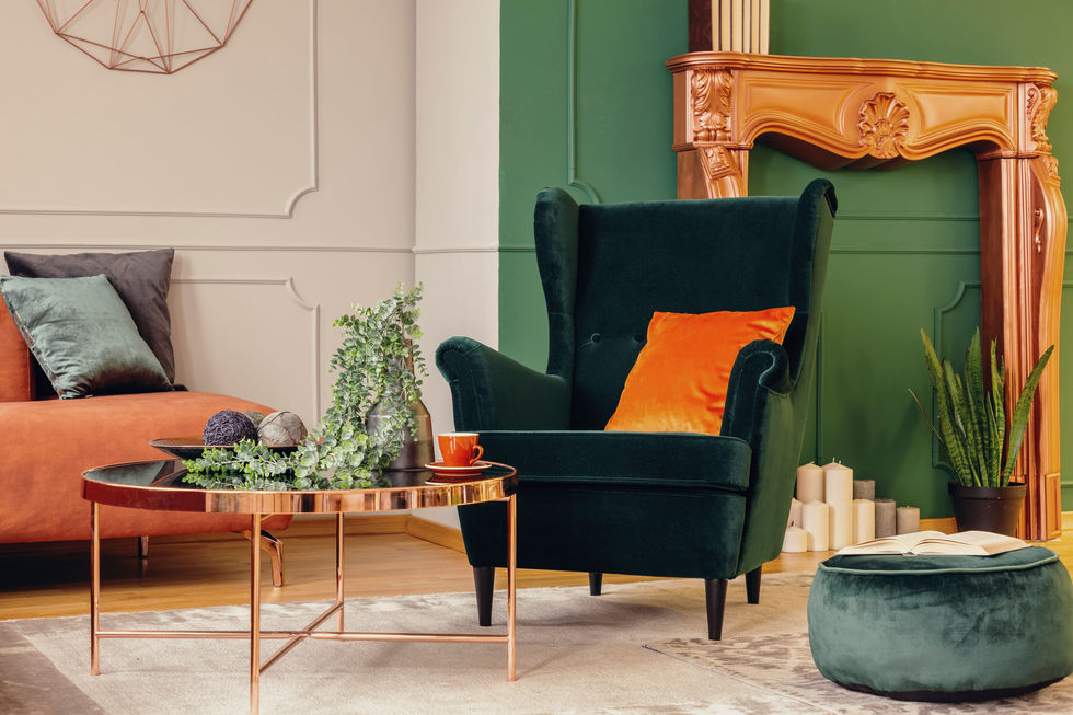 Living room with a dark emerald green velvet wingback armchair, a bright orange throw pillow, a copper coffee table, and a carved wood fireplace mantel