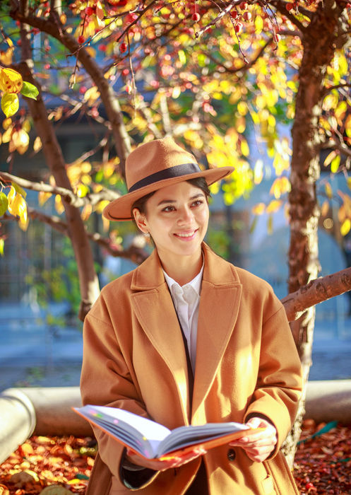 Woman in a camel coat and felt hat reading a book under colorful autumn tree foliage