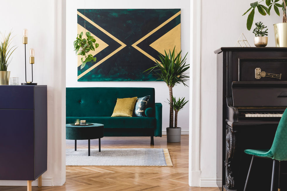 Living room with an emerald green velvet sofa and armchair, a mustard yellow pillow, a dark abstract gold and green painting, a potted palm, and a black piano