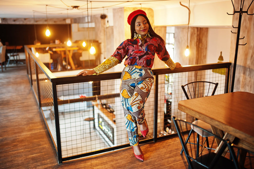 Woman wearing a red beret and a colorful printed blouse and trousers, posing on an industrial railing in a loft-style cafe