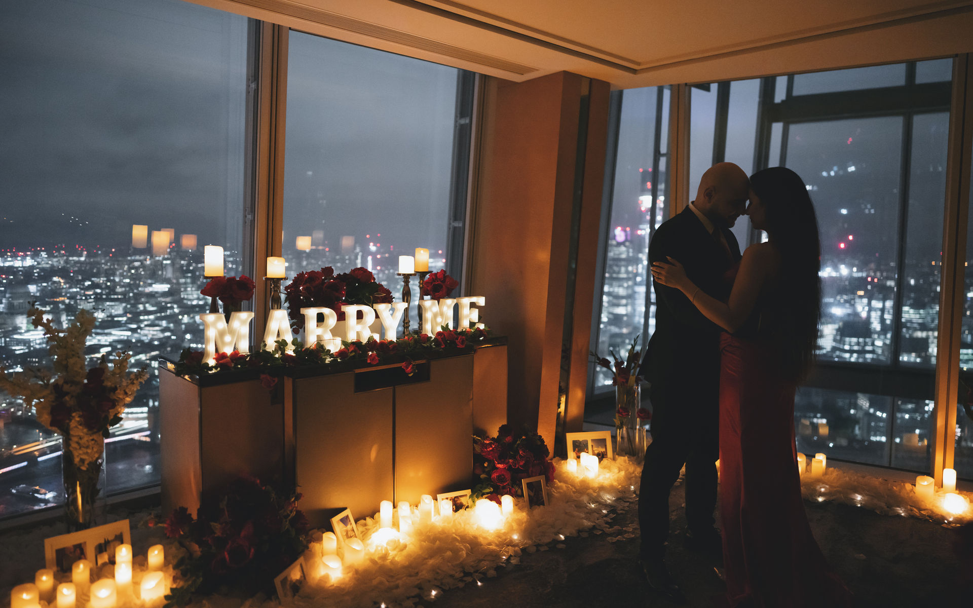 Couple dancing beside illuminated "MARRY ME" sign, proposal setting with city views