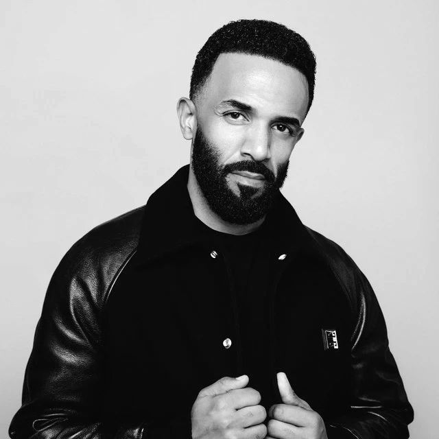 Craig David posing in a black jacket against a neutral background confidently.