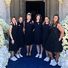 Six women in black dresses standing by a floral arch entrance.