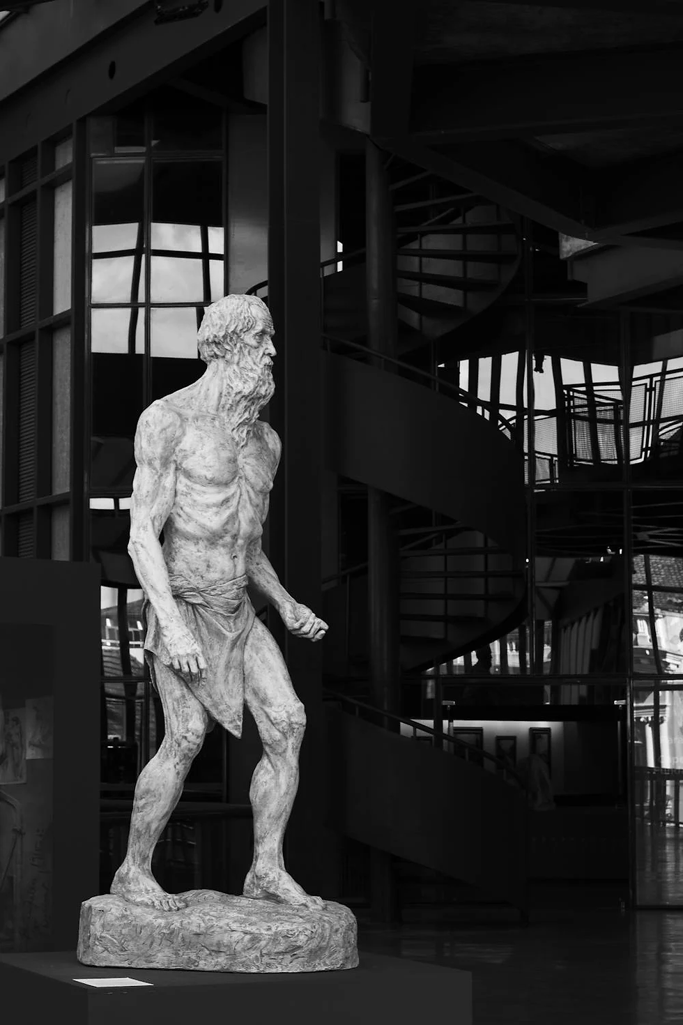 Sculpture of a man, inside a building with a spiral staircase background.