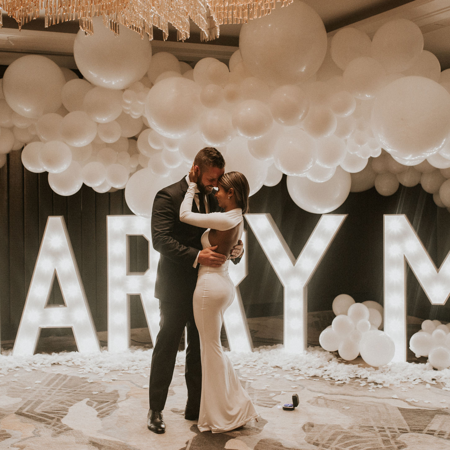 Couple embracing in front of lit-up letters spelling MARRY ME, proposal setting