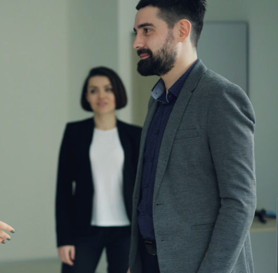 Bearded man in suit smiling, woman in blazer in background