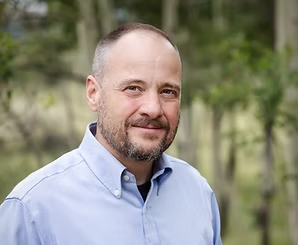 Portrait photograph of Eric Wollard. A man with a short beard, wearing a light blue shirt 