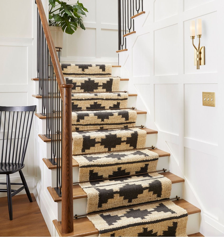 Wooden stairs with a geometric-patterned beige and black runner. White walls, a plant, a wall sconce, and a black chair create a cozy setting.