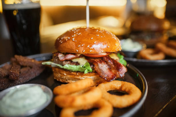 Siyah tabakta domuz pastırmalı hamburger ve soğan halkaları, arka planda bulanık sos kapları.