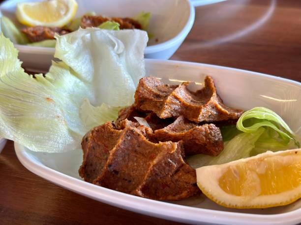 Beyaz tabakta yeşil marul üzerinde çiğ köfte ve limon dilimi. Arka planda ahşap masa ve başka bir tabak görünüyor.