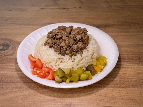 Beyaz plastik tabakta servis edilen pilav üstü et yemeği; etrafında doğranmış domates, yeşil turşu ve sarı turşu garnitürleriyle sunulmuş, yanında kapalı ayran ve sade bir masa düzeniyle görüntülenmiş.