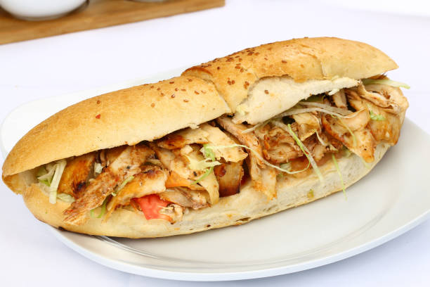 Beyaz tabakta döner ekmek. İçinde tavuk döner, marul ve domates var. Ekmek üzeri susamlı. Arka plan beyaz masa örtüsü.