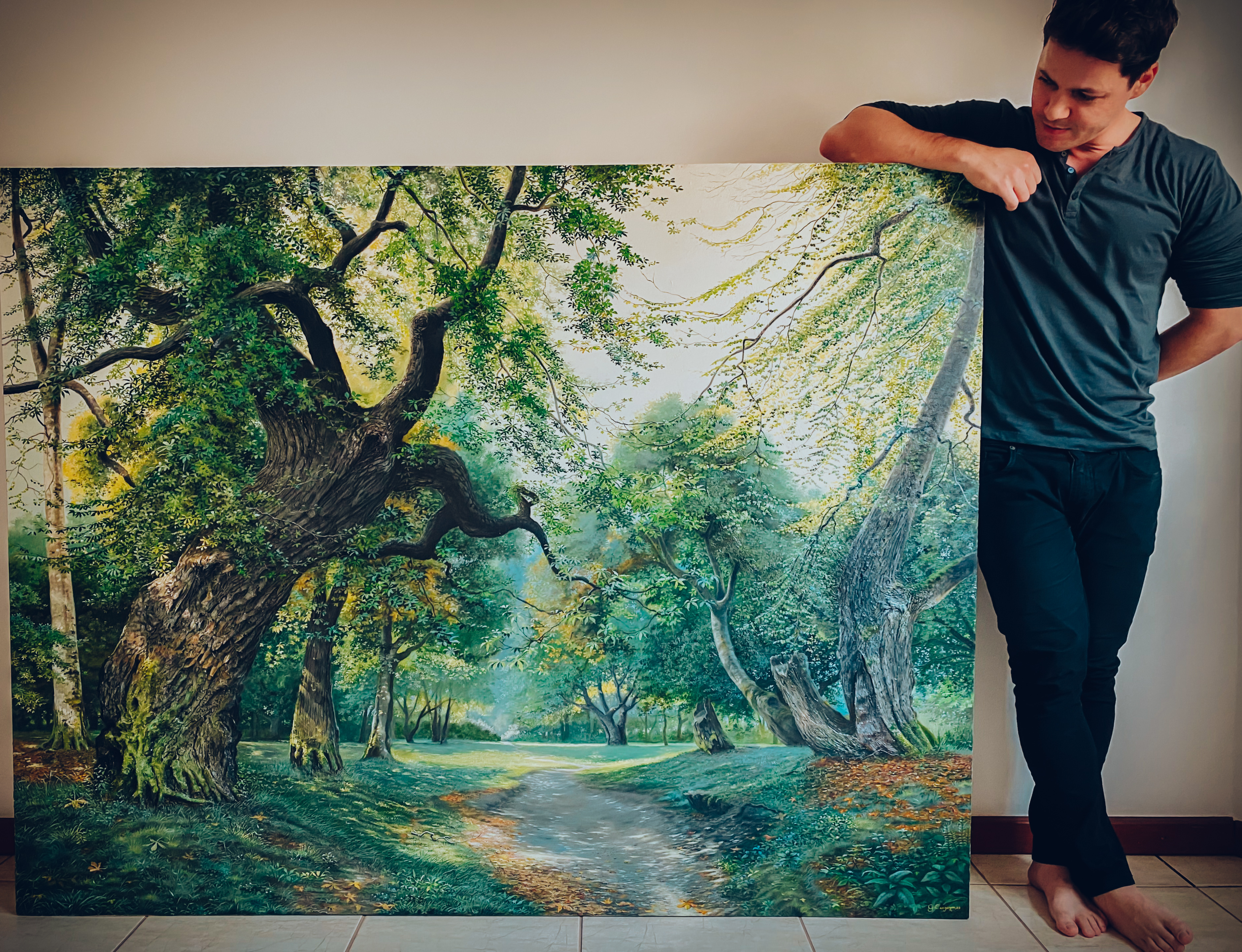 Oil on canvas by artist Gustavo Comparim, who holds his hyper-realistic painting depicting a vibrant forest. The work features twisted, moss-covered trunks and green and golden leaves in an autumnal setting, with stunning detail that conveys texture and depth. The scene captures the grandeur of nature with realism and rich color.