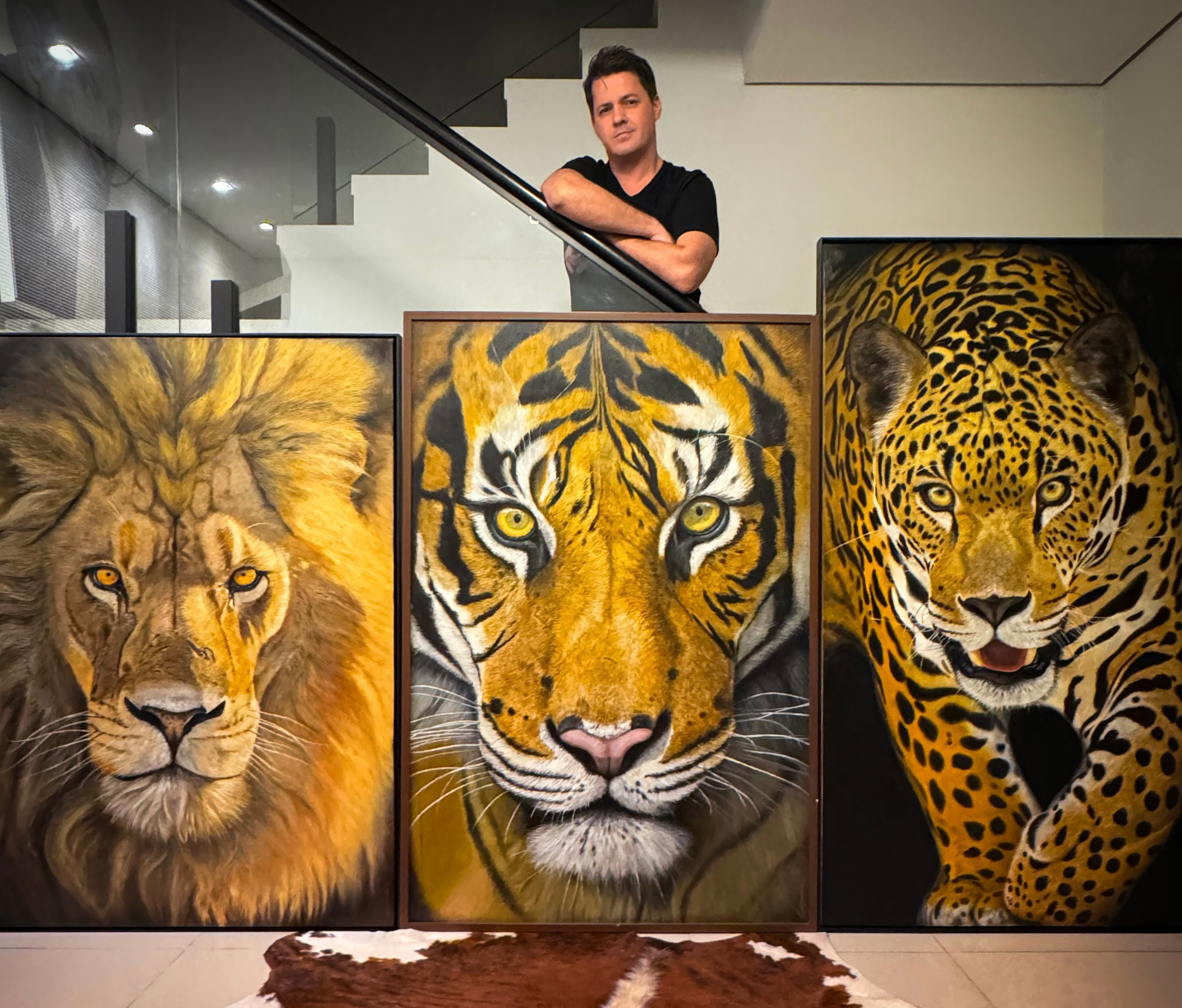 O artista plástico Gustavo Comparim está apoiado no corrimão de uma escada moderna, posicionado acima de três grandes pinturas realistas feitas com a técnica óleo sobre tela. As obras retratam um leão de juba imponente à esquerda, um tigre com olhar penetrante ao centro e uma onça-pintada em posição de ataque à direita. A cena se passa em um ambiente sofisticado, com piso claro e uma grande pele de boi estendida no chão, destacando a imponência das pinturas e a precisão dos detalhes realistas criados pelo artista.