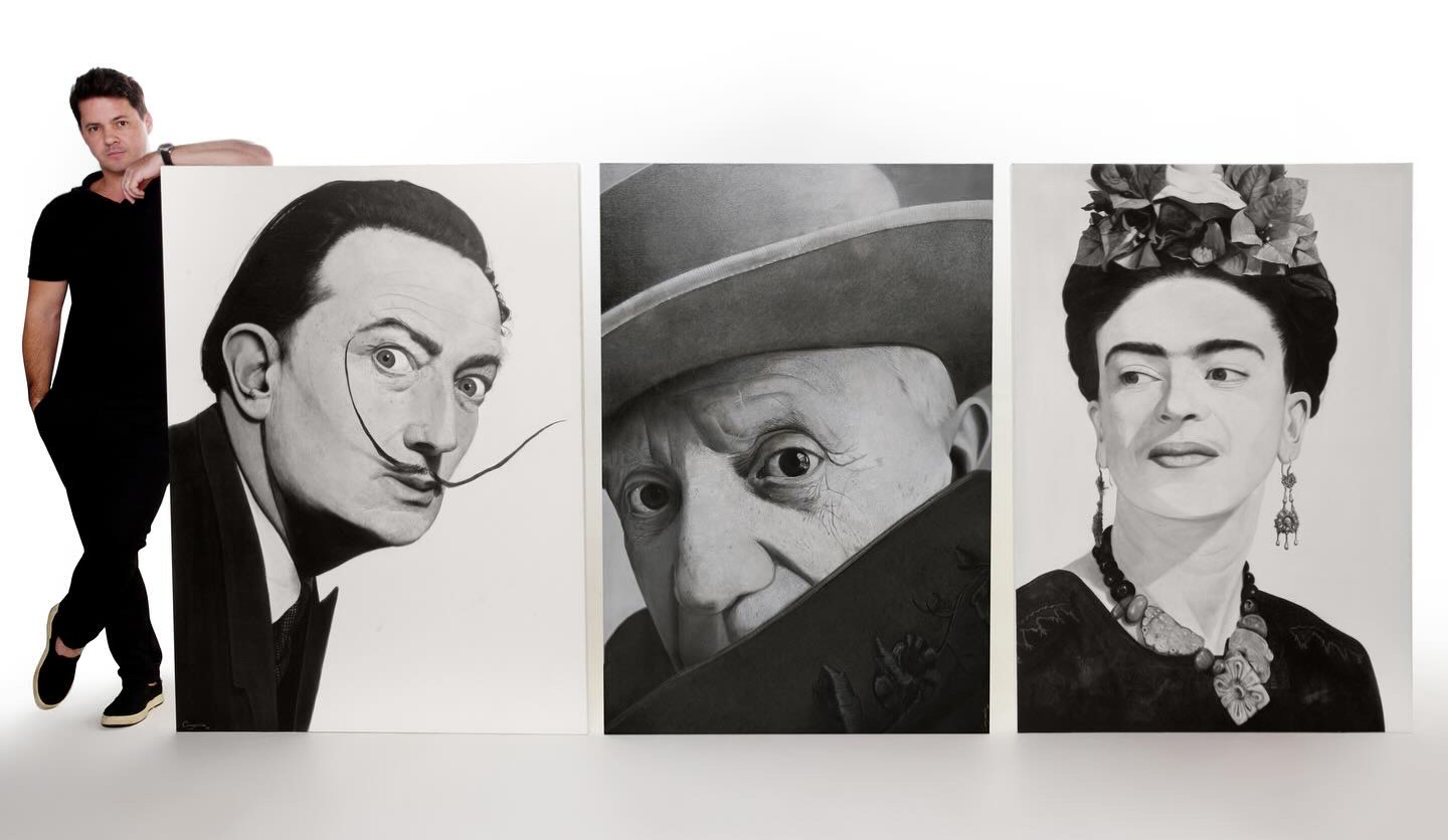 Gustavo Comparim, um homem jovem, branco, de cabelos curtos, vestindo camiseta e calça preta, está em pé ao lado de três pinturas em preto e branco, dispostas no chão contra uma parede branca. As obras retratam, da esquerda para a direita, Salvador Dalí, Pablo Picasso e Frida Kahlo. As pinturas são feitas em óleo sobre tela e possuem traços expressivos, com alto contraste entre luz e sombra, destacando os rostos icônicos de cada artista. O ambiente tem iluminação suave, enfatizando os detalhes das obras.