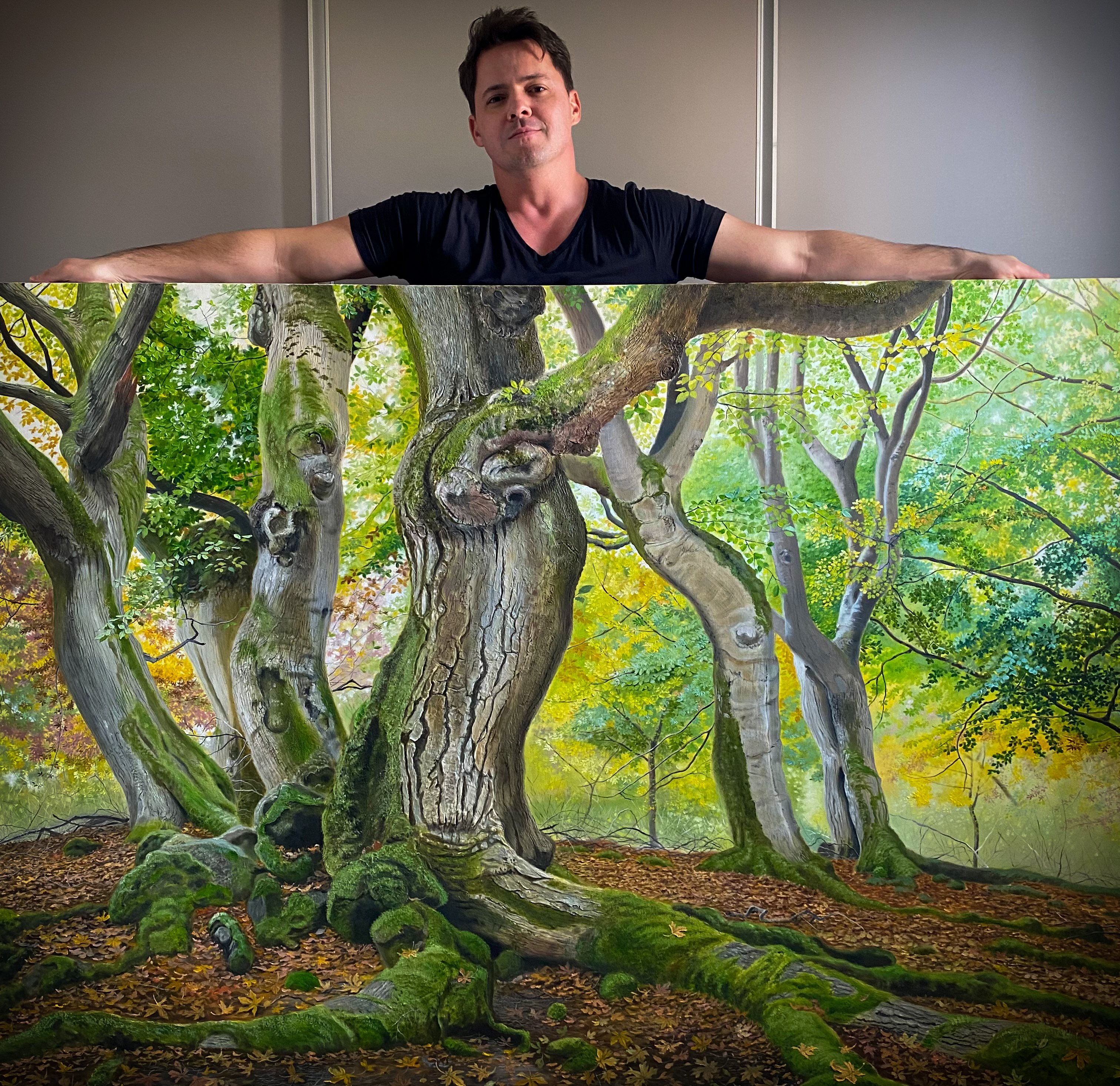 Oil on canvas by artist Gustavo Comparim, who holds his hyper-realistic painting depicting a vibrant forest. The work features twisted, moss-covered trunks and green and golden leaves in an autumnal setting, with stunning detail that conveys texture and depth. The scene captures the grandeur of nature with realism and rich color.