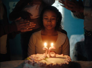 Fotograma de la película Niñas Escarlata. En un ambiente oscuro, una adolescente de piel morena y cabello rizado hasta los hombros está sentada frente a un pastel de cumpleaños blanco, mirando hacia abajo con expresión seria. Dos velas encendidas, con forma de los números «16», están clavadas en el pastel.