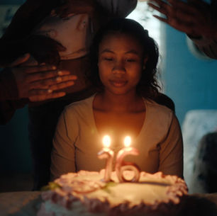 Fotograma de la película Niñas Escarlata. En un ambiente oscuro, una adolescente de piel morena y cabello rizado hasta los hombros está sentada frente a un pastel de cumpleaños blanco, mirando hacia abajo con expresión seria. Dos velas encendidas, con forma de los números «16», están clavadas en el pastel.