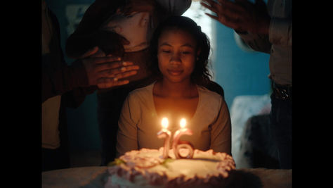 Fotograma de la película Niñas Escarlata. En un ambiente oscuro, una adolescente de piel morena y cabello rizado hasta los hombros está sentada frente a un pastel de cumpleaños blanco, mirando hacia abajo con expresión seria. Dos velas encendidas, con forma de los números «16», están clavadas en el pastel.