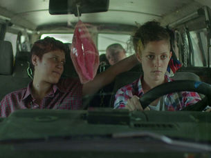 Fotograma de una película que muestra a dos jóvenes dentro de una van. A la derecha, una joven de cabello rizado recogido en lo alto conduce el vehículo, sujetando el volante y mirando al frente con expresión seria. A la izquierda, otra joven, de cabello corto y vestida con una camisa de cuadros roja, mira a la conductora.