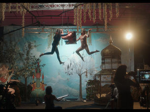 Dos personas bailan suspendidas en el aire en un escenario teatral, rodeadas de plantas, árboles y elementos decorativos, mientras otras personas observan y graban la escena.