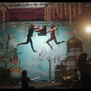 Dos personas bailan suspendidas en el aire en un escenario teatral, rodeadas de plantas, árboles y elementos decorativos, mientras otras personas observan y graban la escena.