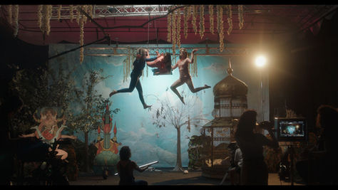 Dos personas bailan suspendidas en el aire en un escenario teatral, rodeadas de plantas, árboles y elementos decorativos, mientras otras personas observan y graban la escena.