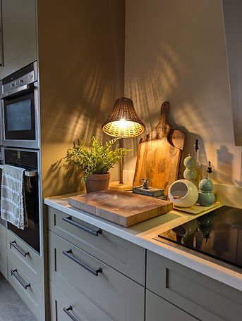 Detail of the refinished Treron kitchen, showing Shaker drawers with bronze hardware, an induction hob, timber chopping boards, greenery and a small warm-glowing lamp beside the integrated ovens.