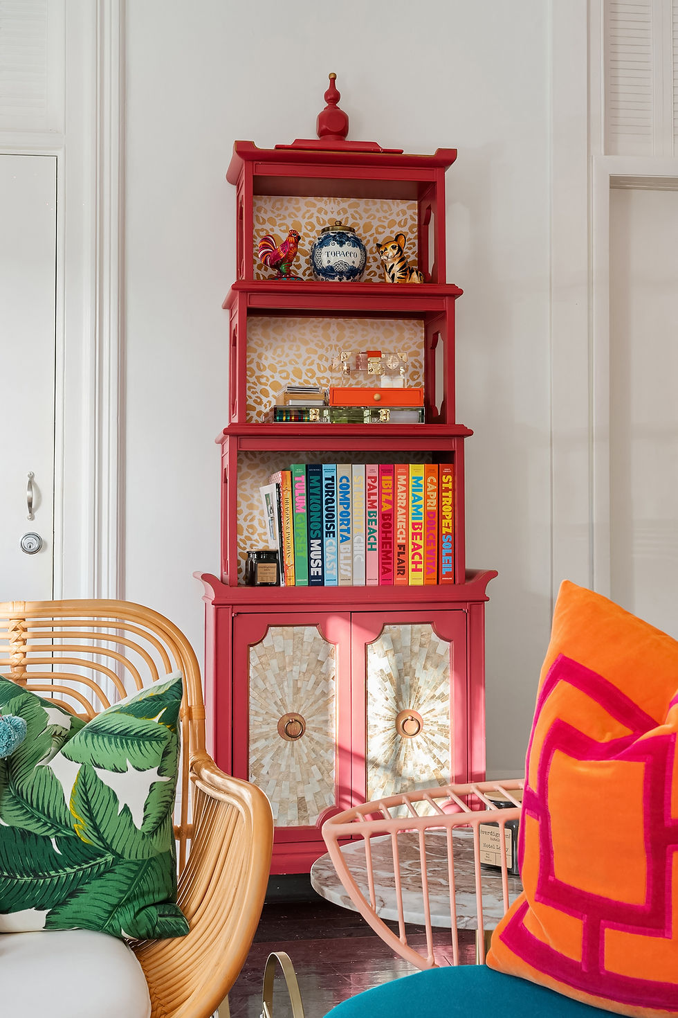 A red lacquer etagere filled with books and objects, beside a rattan chair with palm-print and orange cushions
