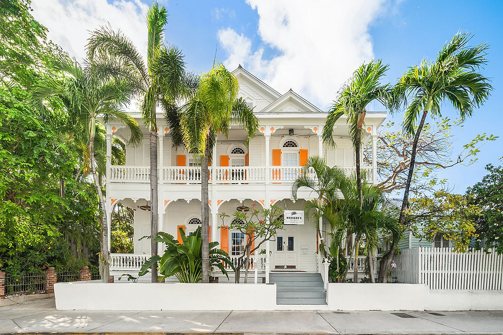 The white Victorian facade of Marrero's Guest Mansion behind palm trees, with orange shutters and a two-storey veranda