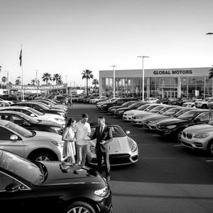 People at a car dealership during a summer sales event. Rows of cars, palm trees, and "GLOBAL MOTORS" sign in the background. Black and white.
