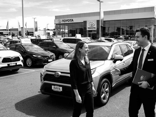 A woman and a man in a car dealership lot smile as they converse beside a Toyota RAV4. The building and multiple cars are in the background.