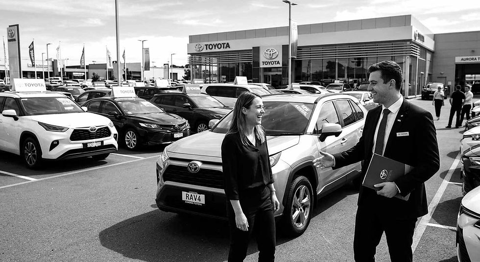 A woman and a man in a car dealership lot smile as they converse beside a Toyota RAV4. The building and multiple cars are in the background.