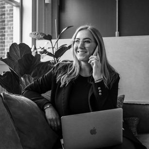 Smiling person on phone with laptop in modern office, seated on a sofa. Large plant and window in background. Black and white image.