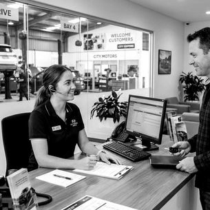 A receptionist smiles at a customer holding keys in a car service centre. Signs read "Service Drive" and "Welcome Customers." Cars in the background.