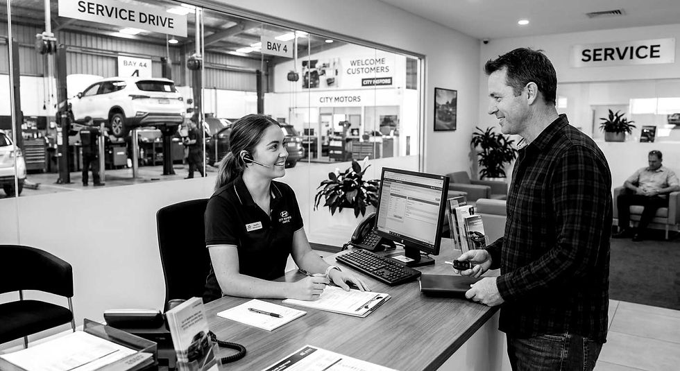 A receptionist smiles at a customer holding keys in a car service centre. Signs read "Service Drive" and "Welcome Customers." Cars in the background.