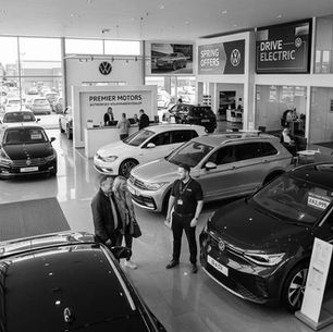 Customers and a salesman in a Volkswagen car showroom. Cars are displayed with signs. Bright, modern interior with large windows.