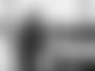 Car showroom with a salesperson in a suit showing a car to a woman. Another pair in background. Large windows, modern setting. Grayscale.