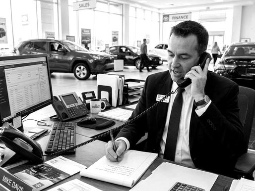 Man in a sales office talks on the phone and writes notes, surrounded by car brochures and a computer. Cars visible in the showroom.