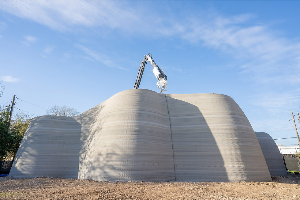 A large 3D printer constructs a curved, layered concrete structure under a clear blue sky. Shadows and light create a textured effect.