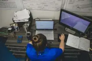 woman leaning over a laptop with a larger monitor next to her. clearly working from home with kids is exhausting
