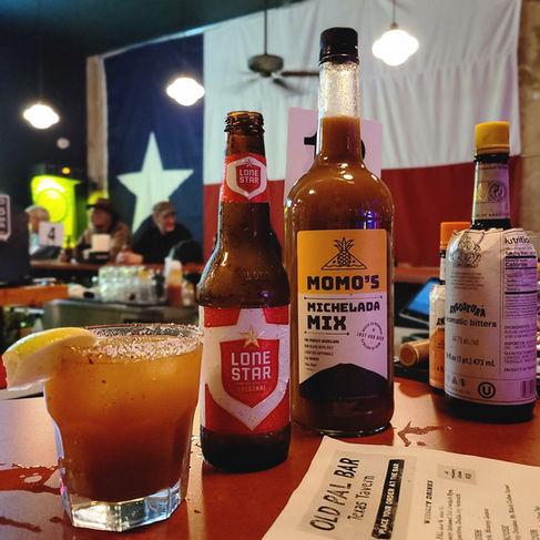 lass of michelada and Momo’s Michelada Mix on table with Texas flag backdrop, bar setting in Austin, TX