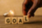 Hand places "L" block to form "GOAL" on wood surface; glowing bulb in background, conveying creativity and aspiration.